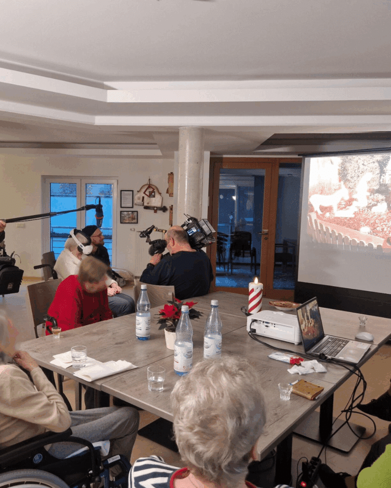 In einem hellen Gemeinschaftsraum sitzen mehrere Menschen an einem Tisch, auf dem Wasserflaschen, Gläser und ein Laptop stehen. Ein Beamer projiziert ein Bild an die Wand, während Kamera- und Tontechnik die Vorführung einer VR-Brille begleitet.